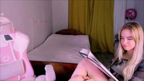 A video of a blonde woman reading a magazine in a dimly lit bedroom with a white bed, yellow curtains, and a pink backpack.