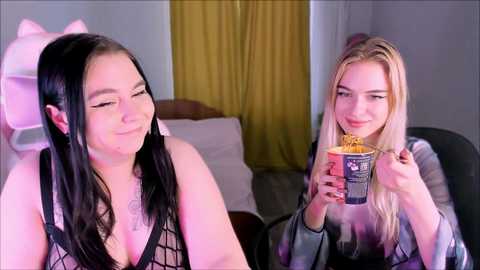 Video of two young women with fair skin, one with long black hair in a black fishnet top, and the other with long blonde hair in a grey sweater, eating popcorn in a room with yellow curtains.