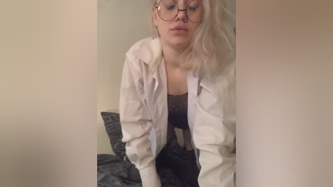 A video of a blonde woman with glasses, wearing a white shirt and dark top, leaning forward, indoors.