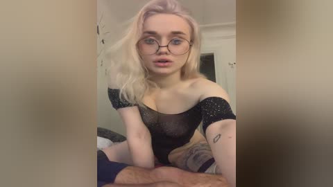 Video of a young woman with light skin, blonde hair, and glasses, wearing a black, glittery off-shoulder top, seated on a man's lap in a dimly lit room.