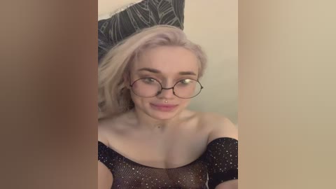 Video of a fair-skinned, blonde woman with shoulder-length hair, wearing round glasses and a black sparkly off-shoulder top, smiling. Background features a dark leaf patterned pillow.