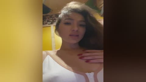 Media: A video of a woman with light skin and long brown hair, wearing a white top, standing against a yellow wall with a hand on her chest.