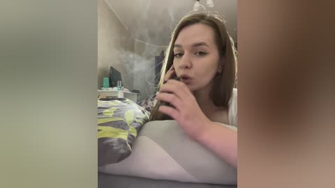 Media: Video of a woman with light skin, light brown hair, wearing a white shirt, smoking a cigarette in a dimly lit bedroom with a messy bed and cluttered nightstand.