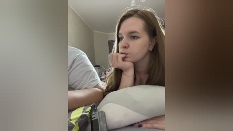 Media: Video of a young Caucasian woman with long brown hair, wearing a light-colored top, lying on her side in bed, resting her head on her hand, with a blurred background.