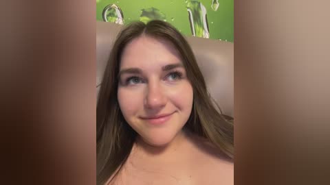 Video of a young, fair-skinned woman with long brown hair, smiling, with a blurred background featuring green and metallic objects.