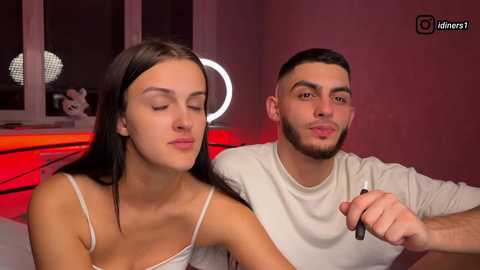 Video of a young Caucasian woman with long brown hair, wearing a white camisole, and a bearded Middle Eastern man in a white t-shirt, holding a vaping device. Background features a red-lit room with a round mirror and plush toys.