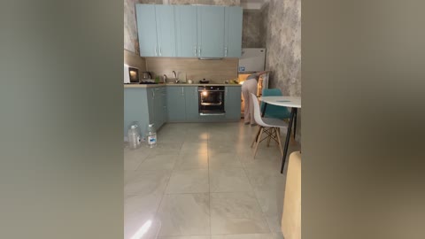 A video of a modern, small kitchen with light blue cabinets, beige countertops, a small oven, and a dining area with a white table and teal chairs. The walls have a rustic, textured finish.