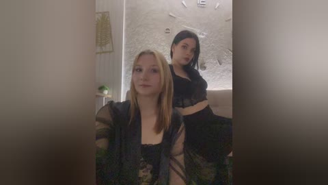 Video of two young women in a bathroom. One, blonde with a sheer black top, stands in the foreground. The other, brunette with long hair, sits behind her in black lingerie.