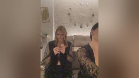 A video shows a blonde woman in a black lace top and skirt, seated on a sofa, hands clasped, in a dimly lit living room with a patterned wall and abstract art.