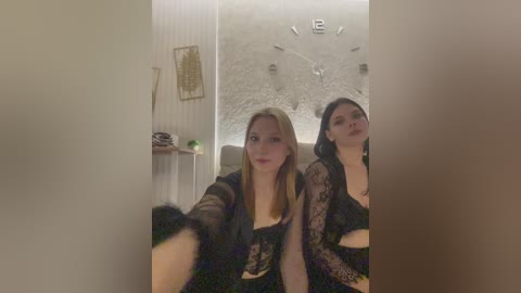Video of two young women with light skin, one blonde, one brunette, wearing black lace lingerie, sitting on a couch in a dimly lit, minimalist room with beige walls and a large clock on the wall.