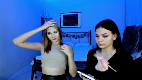 Video of two young women in a dimly-lit bedroom with blue lighting. One is blonde with a beige crop top, touching her head. The other is brunette, applying makeup, with a pink brush.