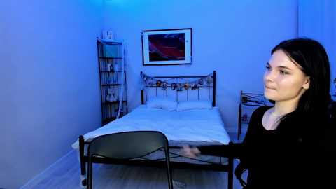 Video of a woman with long black hair, wearing a black sweater, standing in a dimly lit bedroom with a metal bed, blue walls, and modern art on the wall.