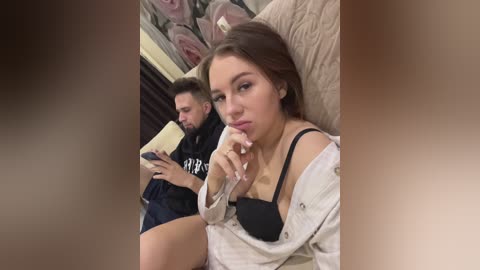 Media: Video of a young woman with light skin, brown hair, and a black bra, sitting on a beige couch, looking at a smartphone. A man in a black hoodie sits behind her, also on the couch, reading a book.
