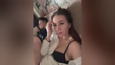 A video of a young woman with light skin, brown hair, and a black bra, lying on a beige sofa next to a man with a beard, wearing a black shirt.
