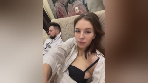 Video of a young Caucasian woman with light skin, brown hair, and a slender physique, partially undressed in a black bra, reclining on a beige couch. Behind her, a man with short dark hair and a beard is also partially undressed.