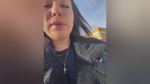 Video of a woman with medium skin tone, long dark hair, wearing a black jacket and silver cross necklace, outdoors under a clear sky with a yellow building in the background.