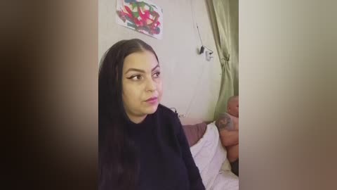 Video of a young Latina woman with long, straight black hair, wearing a black robe, sitting on a bed with a green curtain. Her expression is neutral. A colorful painting hangs on the wall behind her.