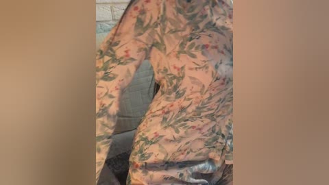 Media: Video of a person wearing a long-sleeved shirt with a floral pattern, featuring green leaves and pink flowers, partially covering their face. Background includes a beige wall and a grey couch.