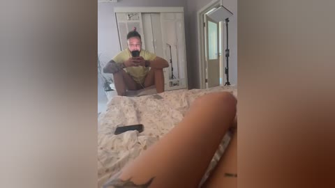 Media: A video of a man sitting on a bed, taking a selfie in a mirror, wearing a yellow shirt and shorts. The room has a white wardrobe and a door.