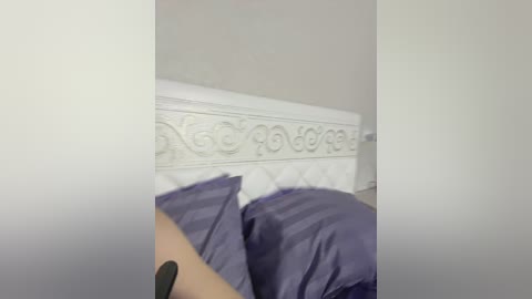 A video of a bed with a white, ornately carved headboard and purple pillows, partially covered by a hand holding a black phone. The walls are light grey, creating a serene, minimalist bedroom ambiance.