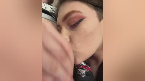 Media: Video of a fair-skinned woman with freckles, closed eyes, and dramatic winged eyeliner, kissing a man's bare chest. She wears a floral top, and his skin tone contrasts with hers.