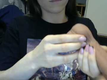 A video of a young woman with shoulder-length dark hair, wearing a black T-shirt with a graphic design. She is holding her own hand in front of her face, with light pink nails. The background features a blurred, dark-colored room with a wooden shelf visible.
