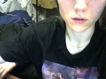 Video of a fair-skinned, blonde-haired woman with a light complexion, wearing a black T-shirt with a graphic design. She has a neutral expression and is positioned indoors with clothes hanging behind her.