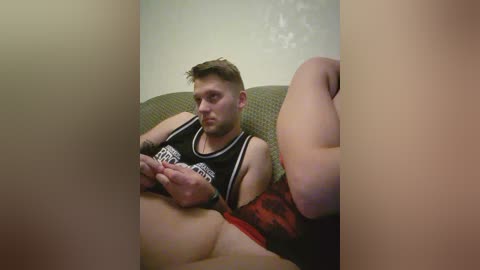Video of a fair-skinned, muscular man with short hair wearing a black sports jersey and red shorts, seated on a green sofa. He is playing with a remote control.