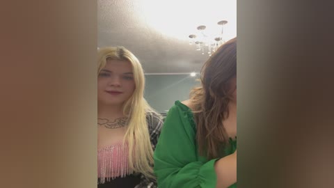 Video of two young women in a dimly-lit room. One with long blonde hair, pink fringed top, black jacket, and necklace. The other has long brown hair and wears a green top.