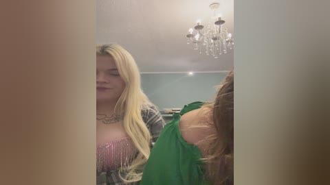 Video of a young woman with long blonde hair, wearing a sparkly pink top and gray cardigan, standing in a teal-painted room with a chandelier.