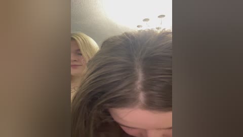 Video of two young women with long, straight, blonde hair, one with a high ponytail, the other with a low ponytail, standing in a dimly lit bathroom with a white ceiling and wall-mounted lights.