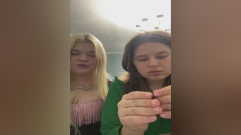 Video of two young women with fair skin. One with long blonde hair, pink top, and a heart tattoo; the other with long dark hair, green top, and a neutral expression. Background shows a ceiling light fixture.