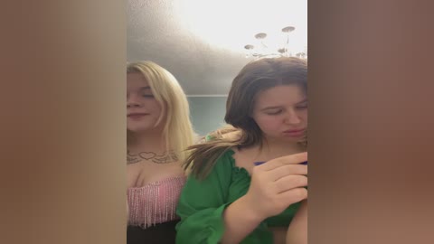 Video of two young women in a dimly lit room; one with blonde hair, pink fringed top, and heart tattoos; the other with brunette hair, green top, and sad expression.
