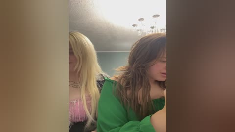 Video of two young women, one with long blonde hair and a pink fringed top, the other with brown hair and a green top, seen from the side, indoors with a chandelier.