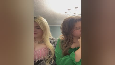 Video of two young women, one with long blonde hair in pink top, the other with brown hair in green shirt, seated side by side, slightly obscured by a vertical divider.