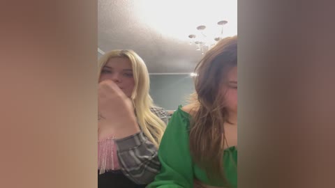 Video of two young women, one with blonde hair and another with brown hair, wearing green and gray clothes, respectively, standing in a dimly lit hallway with chandelier lights.