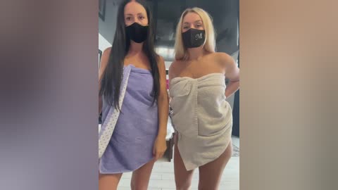 Media: Video of two women, one with long black hair, the other with long blonde hair, both wearing black masks and wrapped in towels, standing in a modern indoor space.