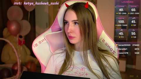 Media: Video of a light-skinned woman with long brown hair and devil horns, wearing a white \"KOTOR\" T-shirt, sitting in a pink gaming chair. Background shows party decorations, and streaming stats on screen.