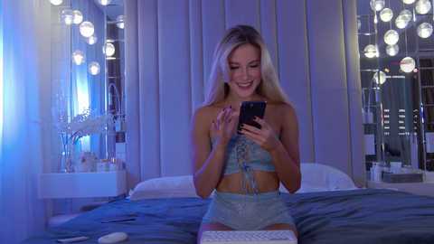 Video of a blonde woman in a blue lingerie set, smiling, holding a phone, in a modern, dimly-lit bedroom with mirrored walls and white bedding.