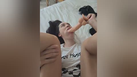 A video of a young woman with pale skin and dark hair lying on a bed, wearing a \"Free Hugs\" t-shirt, holding a large, realistic dildo.