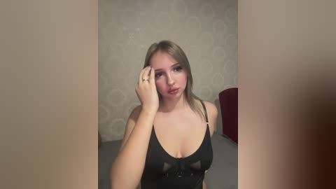 Media: Video of a young, fair-skinned woman with straight, shoulder-length blonde hair, wearing a black lace bralette, touching her face. She stands in a simple room with gray wallpaper and a red chair in the background.