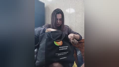 Media: Video of a woman with long brown hair, wearing a black jacket, sitting in a stall. She holds a black bag with a green logo and white text. Background features beige and brown tiled walls.