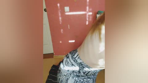 A video shows a partially blurred woman with long blonde hair, wearing a patterned dress, standing in a room with a pink wall and wooden floor.