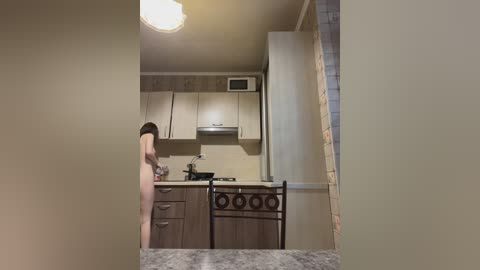 Media: Video of a nude woman with medium breasts, brown hair, standing in a small, modern kitchen with beige cabinets, stainless steel appliances, and a tiled backsplash.