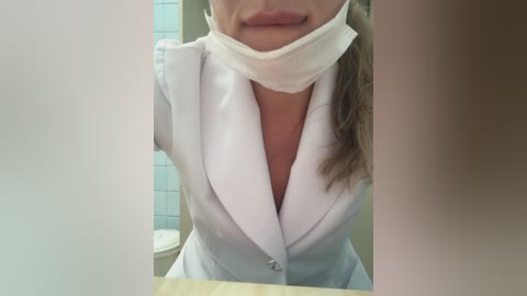 Media: Video of a woman wearing a white surgical mask, light blue medical jacket, and no top underneath, revealing cleavage. Background features a tiled bathroom wall and a toilet.