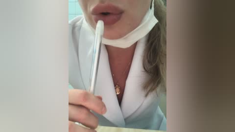 Media: Video of a woman in a white lab coat, wearing a white face mask, brushing her teeth with a white toothbrush. The background is out of focus, highlighting the dental hygiene activity.