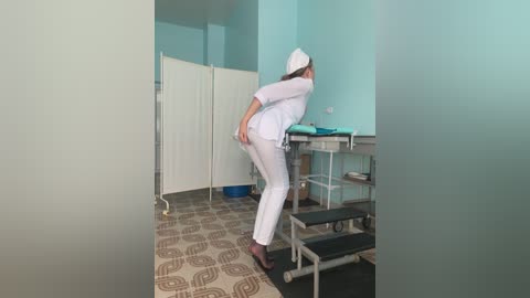 Media: Video of a woman in white medical scrubs bending over a gurney in a clinical room with light blue walls and patterned floor.