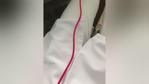 Media: Video of a white, crumpled bedsheet with a red electrical cord running through it, revealing a beige carpet underneath. The image focuses on the bedsheet, highlighting the texture and creases.