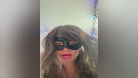 Media: Video of a woman with long, wavy brown hair wearing a black mask with eye slits, pink lipstick, and a blurred background featuring multicolored string lights.