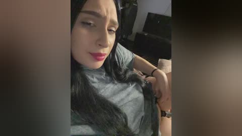 Video of a light-skinned woman with long black hair, red lipstick, and a gray shirt, adjusting her bra. Background includes a TV and dark furniture, indoors.